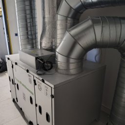 LECH KRUPIŃSKI - Szara centrala rekuperacyjna Dan-Poltherm z podłączonymi, metalowymi rurami wentylacyjnymi w pomieszczeniu. Widoczne naklejki informacyjne na obudowie.