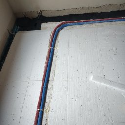 LECH KRUPIŃSKI - Czerwone i niebieskie rury instalacji wodnej lub grzewczej, połączone opaską, ułożone na białych płytach izolacyjnych podłogi w rogu pomieszczenia. Widoczna surowa ściana, czarna membrana i stopa...