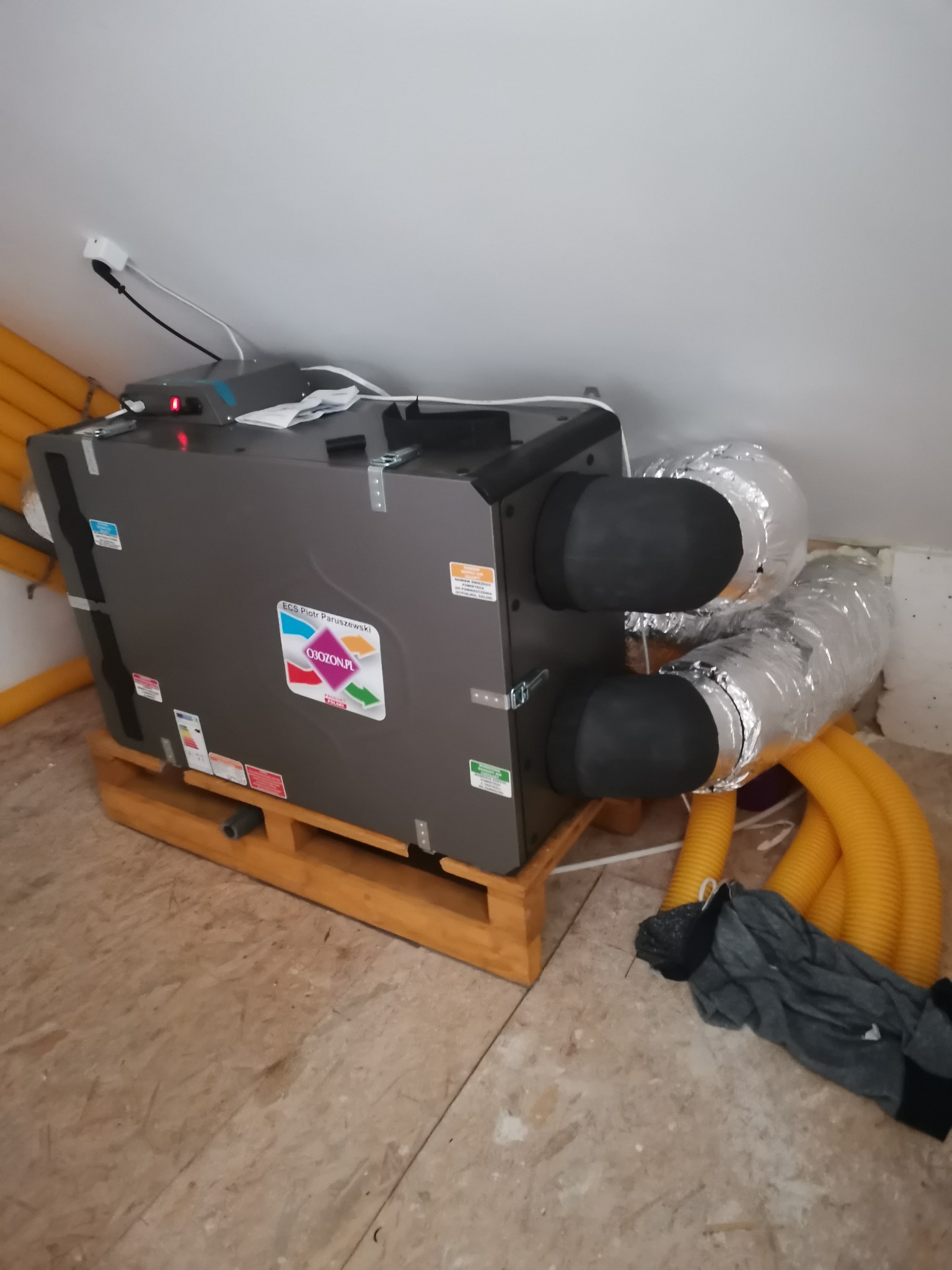 Szara centrala wentylacyjna z podłączonymi przewodami wentylacyjnymi, umieszczona na drewnianej palecie w pomieszczeniu z surowym wykończeniem poddasza.