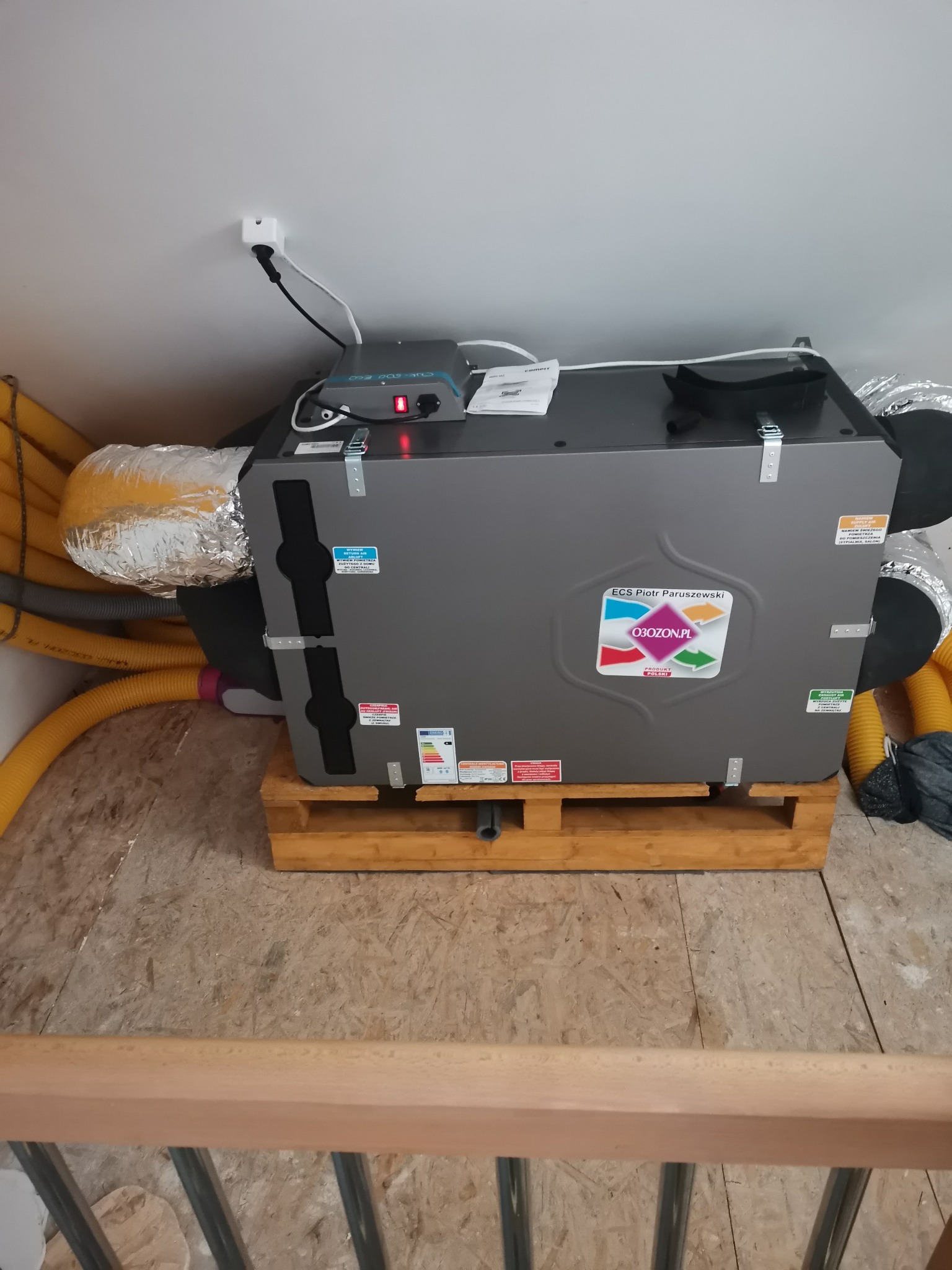 Szara centrala wentylacyjna z podłączonymi przewodami wentylacyjnymi, umieszczona na drewnianej palecie w pomieszczeniu z płyt OSB.