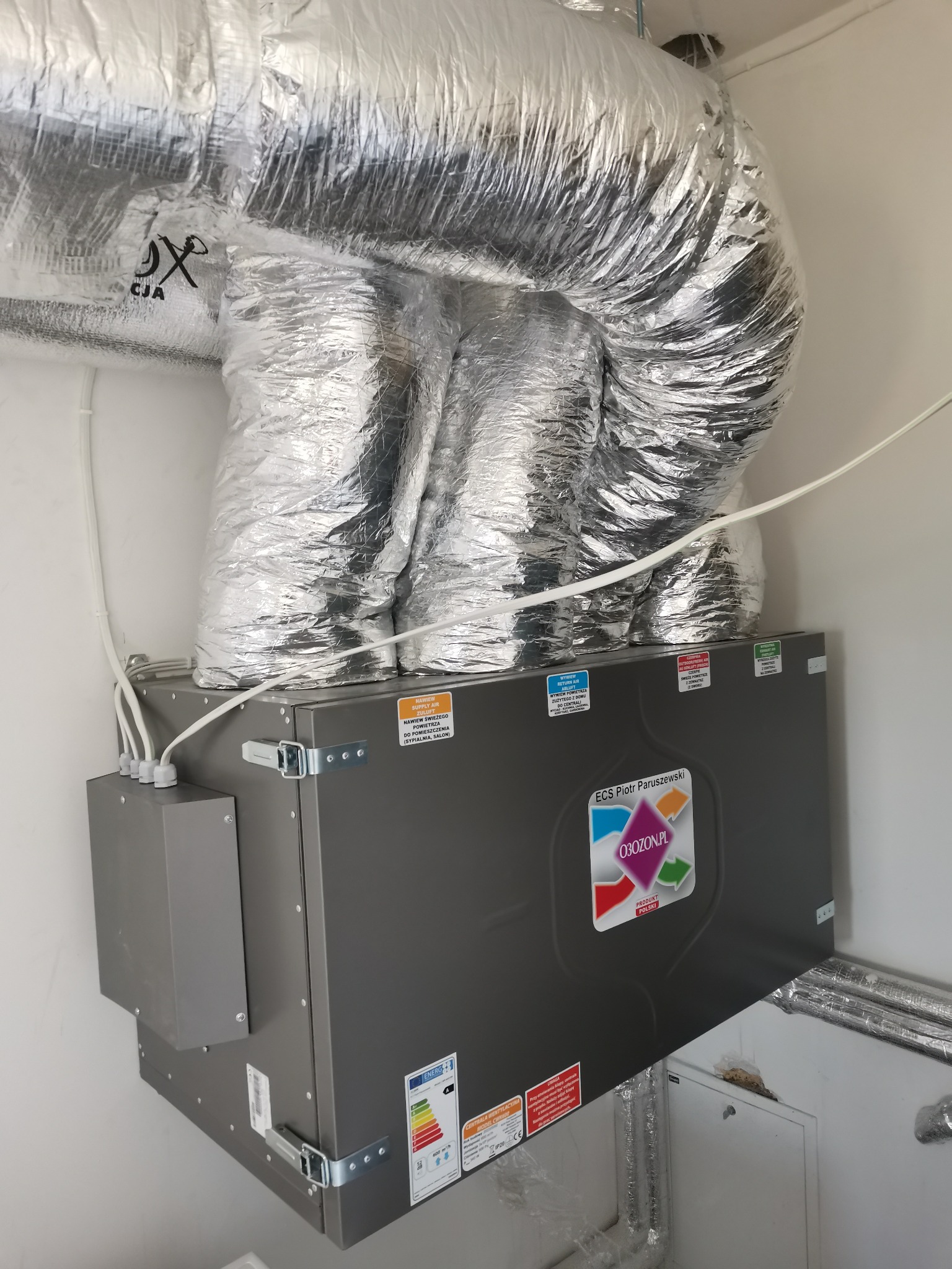Szara centrala wentylacyjna z aluminiowymi przewodami wentylacyjnymi zamontowana przy białej ścianie, widoczne naklejki informacyjne i logo producenta.