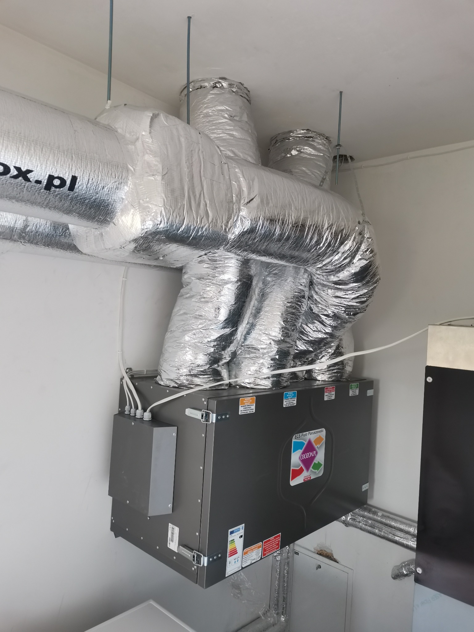 Szare urządzenie wentylacyjne z podłączonymi przewodami izolowanymi folią aluminiową, zawieszone pod sufitem na metalowych prętach w pomieszczeniu z białymi ścianami.