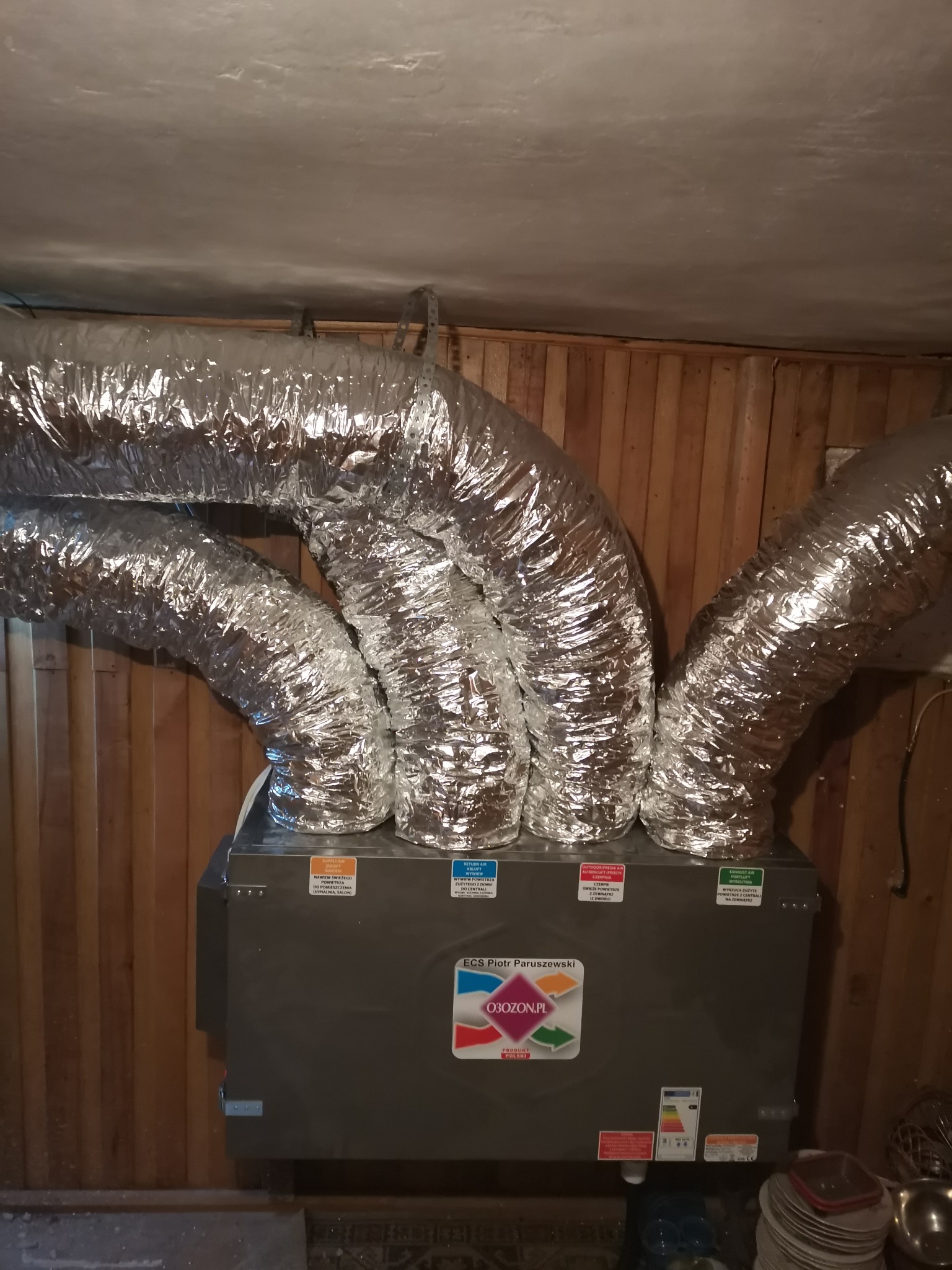 Centrala wentylacyjna z trzema elastycznymi, aluminiowymi przewodami wentylacyjnymi w pomieszczeniu wyłożonym boazerią. Na urządzeniu naklejki informacyjne i logo firmy O3OZON.PL. Widoczne naczynia...