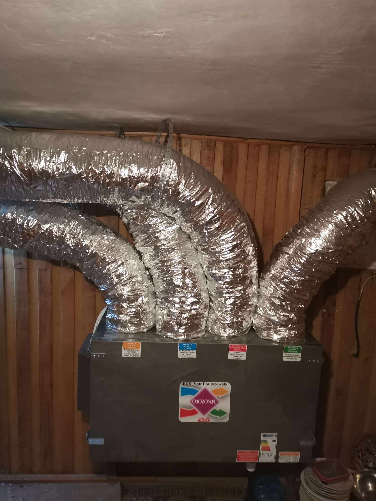 Szare urządzenie rekuperacyjne z czterema podłączonymi elastycznymi, aluminiowanymi przewodami wentylacyjnymi w pomieszczeniu z boazerią.