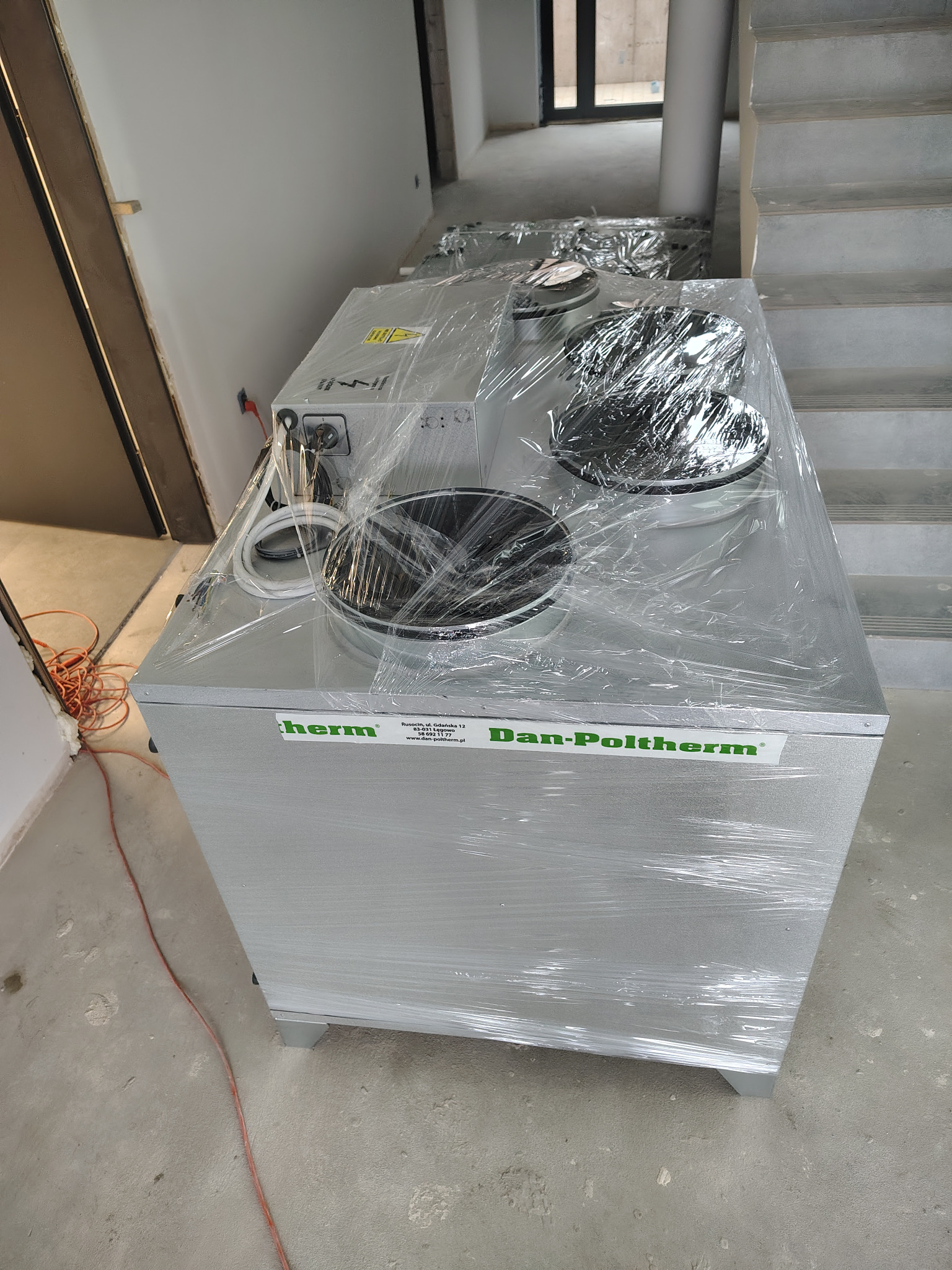 Nowy rekuperator Dan-Poltherm, zabezpieczony folią, ustawiony na podłodze w surowym wnętrzu. Widoczne schody i przygotowane ściany.