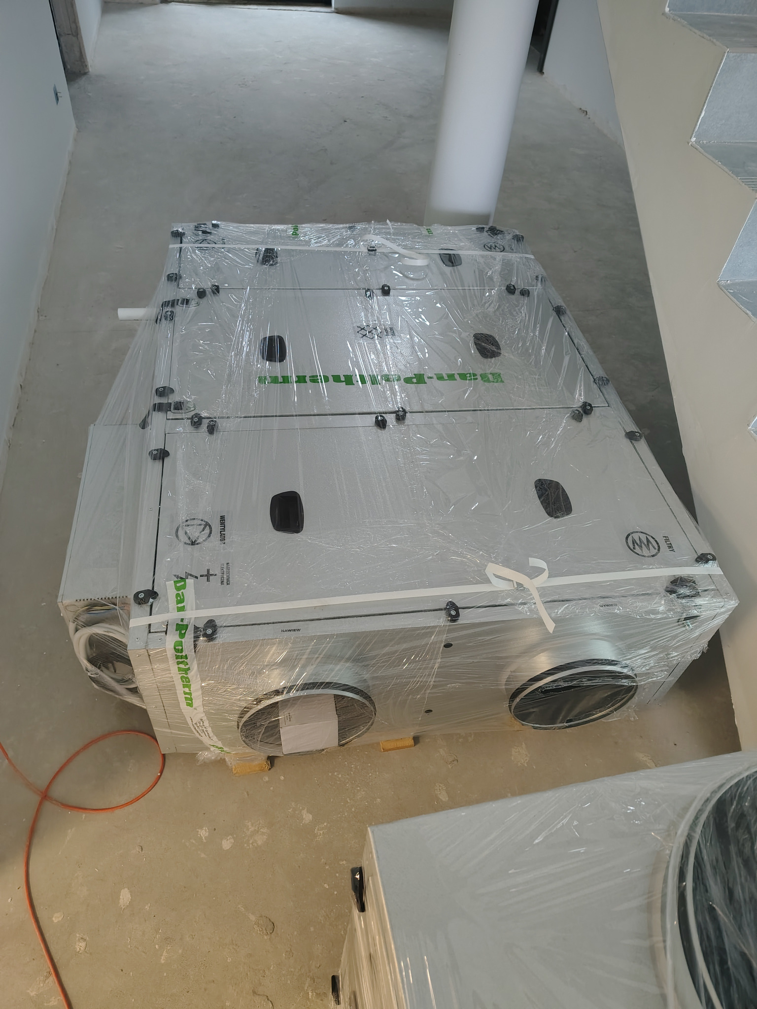 Kompaktowa centrala rekuperacyjna Dan-Poltherm, zabezpieczona folią, czeka na montaż w surowym wnętrzu. Widoczne okrągłe otwory przyłączeniowe i etykiety producenta.