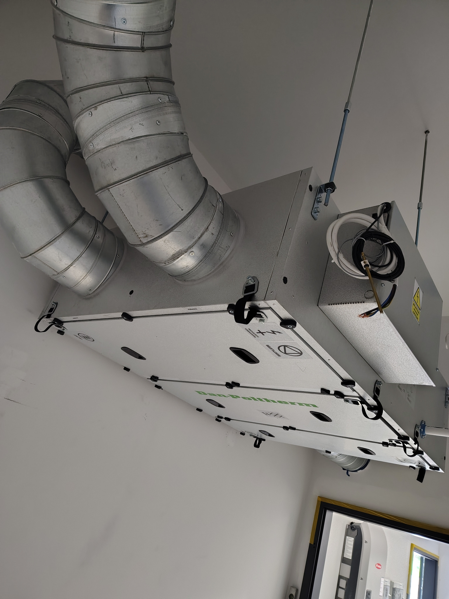 Zamontowany rekuperator Dan-Poltherm na suficie z podłączonymi przewodami wentylacyjnymi i elementami mocującymi. Widok z dołu.