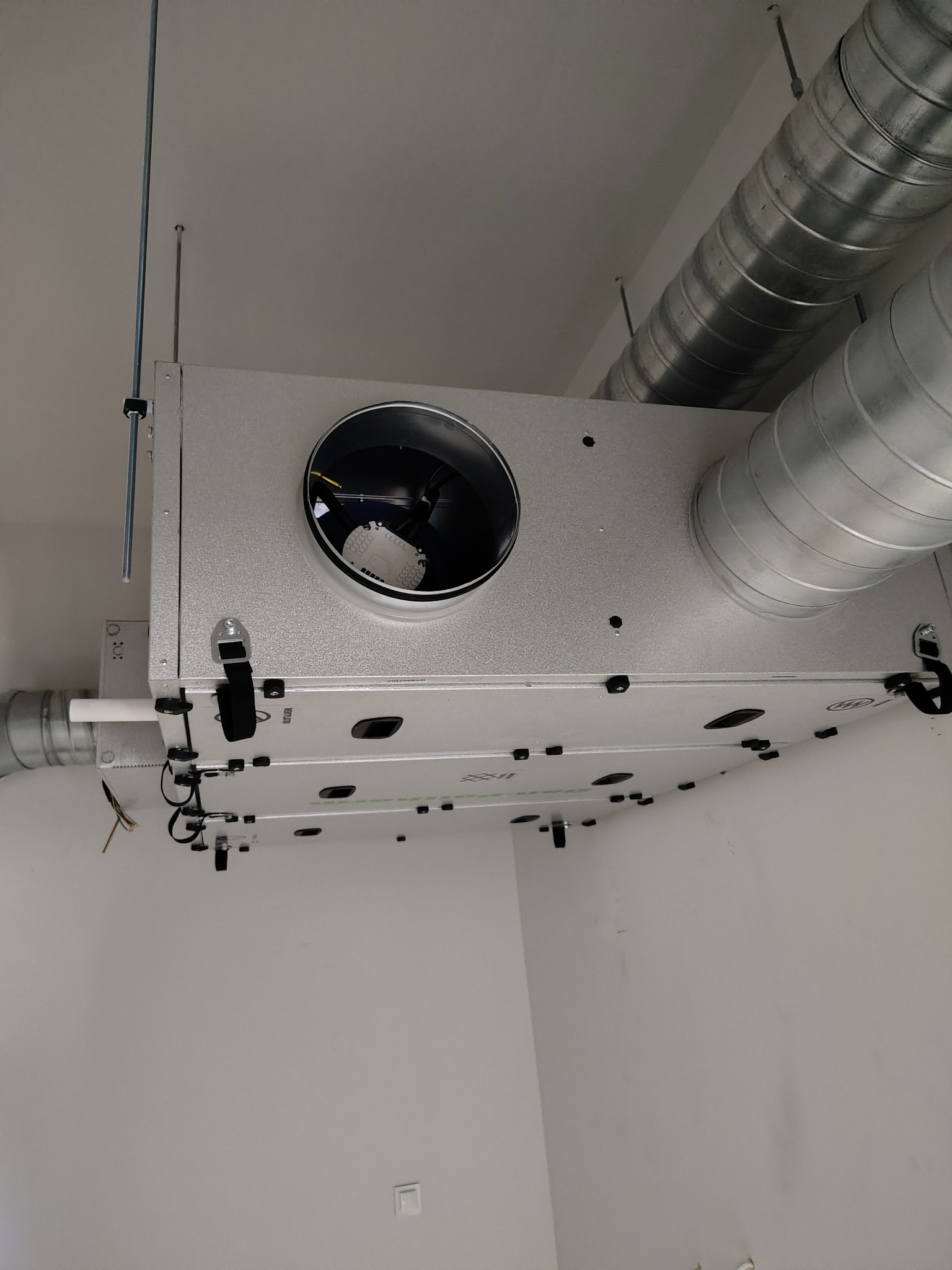 Zamontowany rekuperator pod sufitem z widocznymi kanałami wentylacyjnymi. Srebrna obudowa, czarne elementy montażowe i białe ściany. Precyzyjny montaż instalacji.