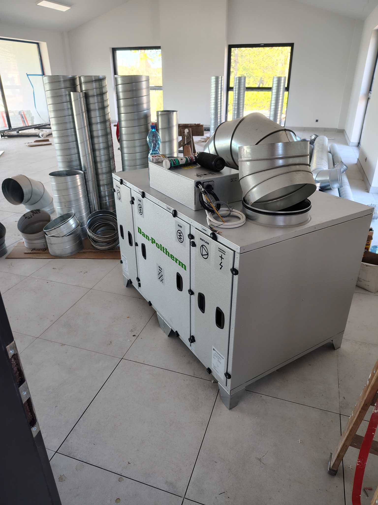 Szary rekuperator Dan-Poltherm z metalowymi rurami wentylacyjnymi w jasnym wnętrzu, przygotowany do montażu. Widoczne okna i drabina.