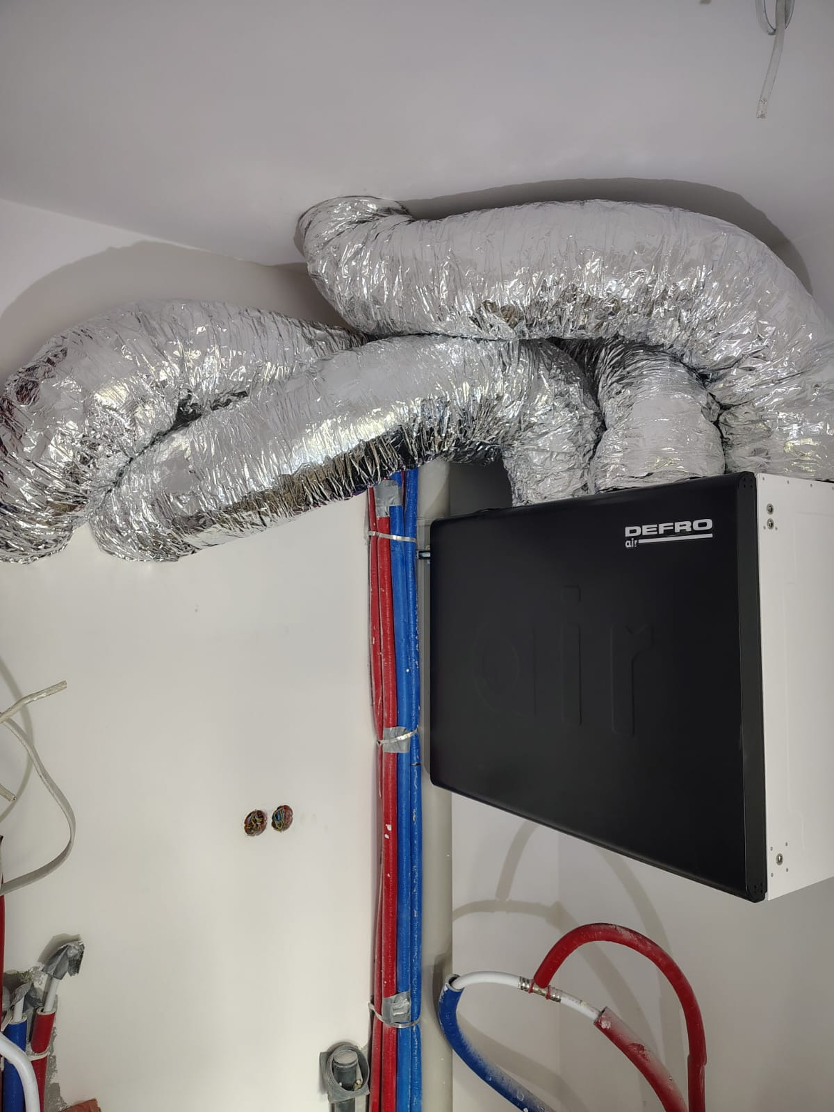 Zamontowany rekuperator Defro Air z widocznymi przewodami wentylacyjnymi i instalacją hydrauliczną w kolorach niebieskim i czerwonym, przymocowaną do ściany.