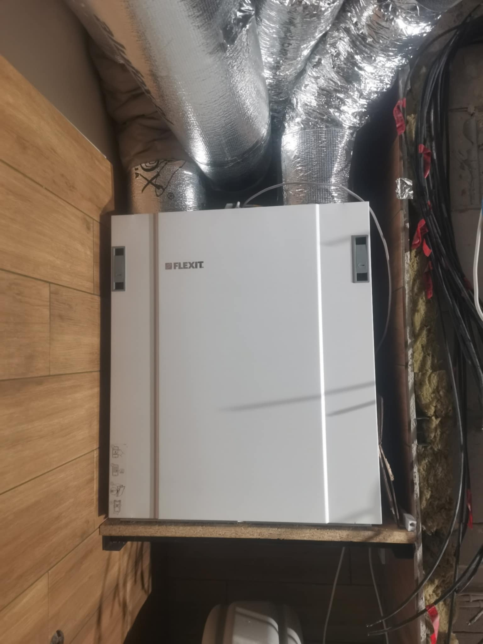 Biała jednostka rekuperacyjna Flexit zamontowana w pomieszczeniu technicznym, widoczne przewody wentylacyjne i instalacje. Ujęcie z bliska, z boku drewniana ściana.