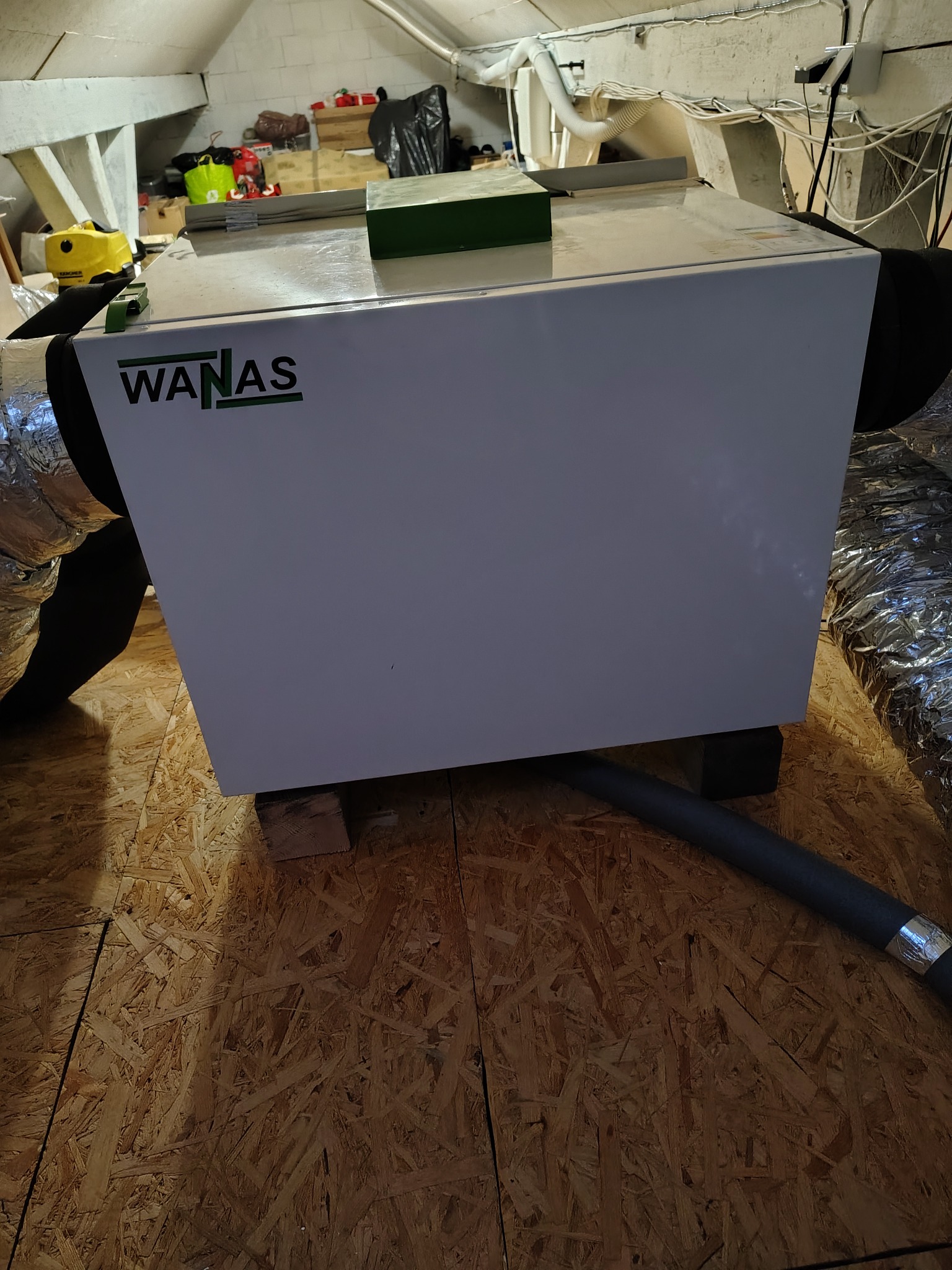 Biała jednostka rekuperacyjna WANAS w przestrzeni poddasza, z widocznymi przewodami wentylacyjnymi i elementami instalacji. Ustawiona na drewnianych klockach.