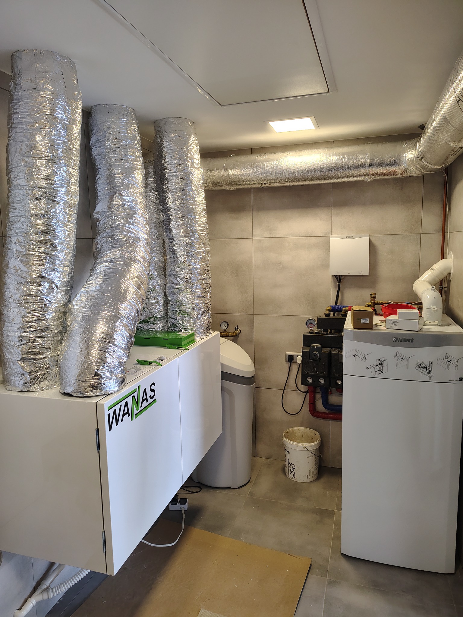 Zmontowana instalacja rekuperacji WANAS z izolowanymi przewodami wentylacyjnymi w pomieszczeniu z szarą glazurą. Widoczny również bojler Vaillant i zmiękczacz wody.