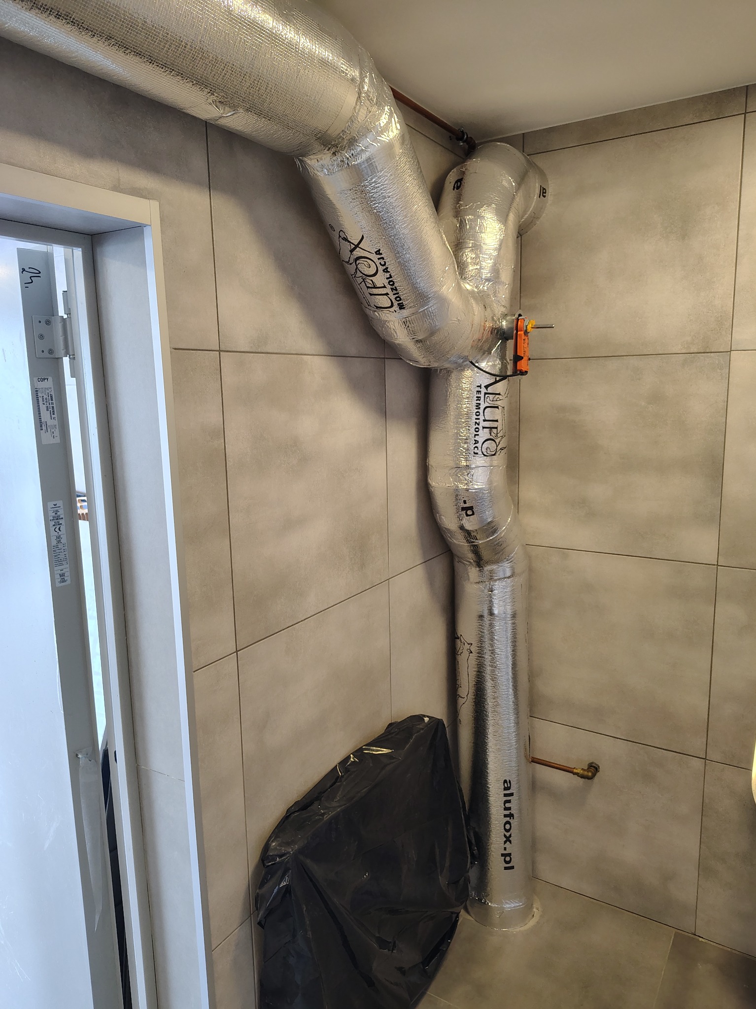 Szczegół instalacji wentylacyjnej z izolacją termiczną Alufox, widoczny fragment rury miedzianej i czarny worek na podłodze, na tle płytek ceramicznych.