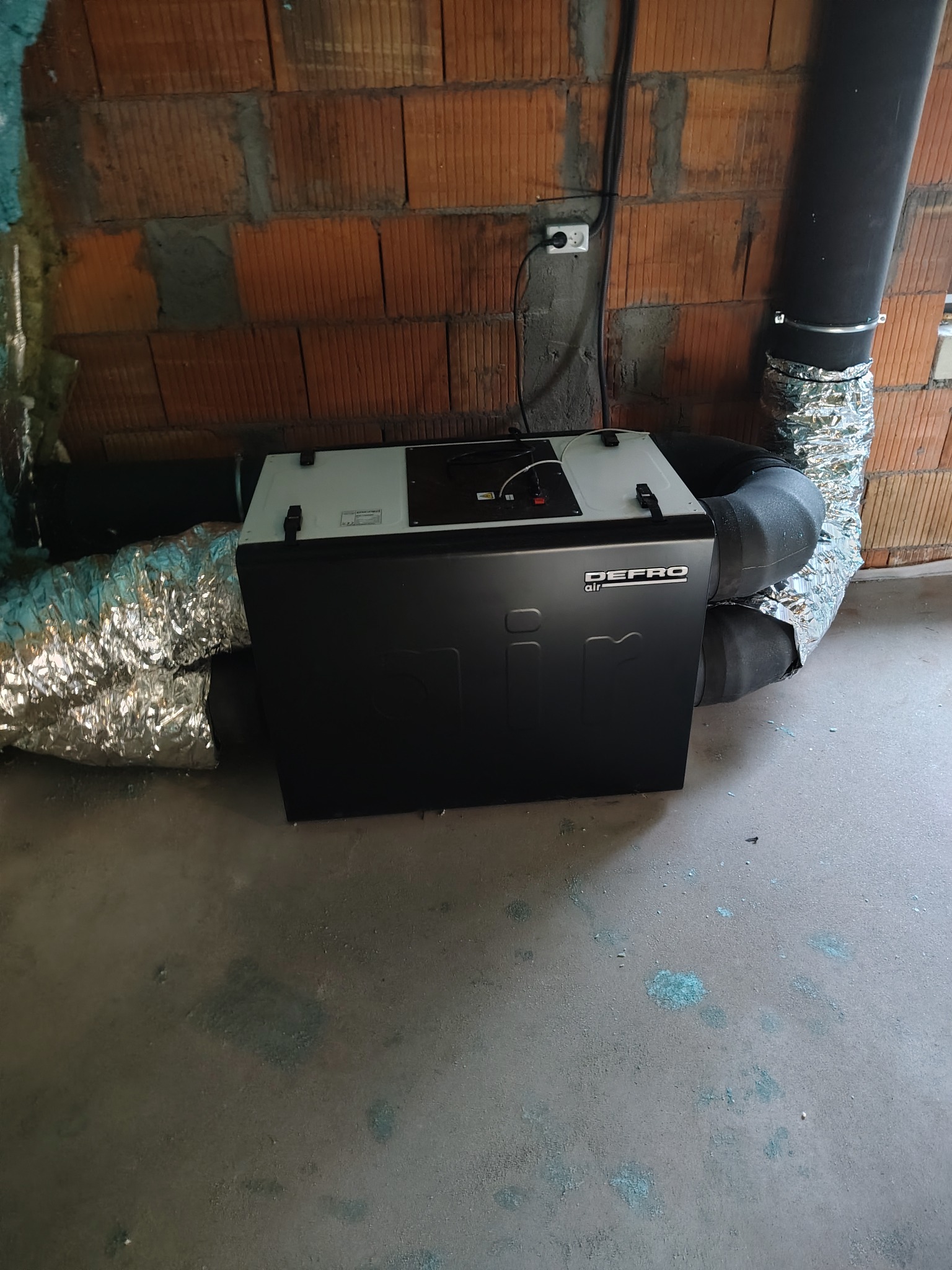 Czarna jednostka rekuperacyjna Defro Air z podłączonymi przewodami wentylacyjnymi na tle ceglanej ściany i surowej podłogi. Widoczne zasilanie elektryczne.