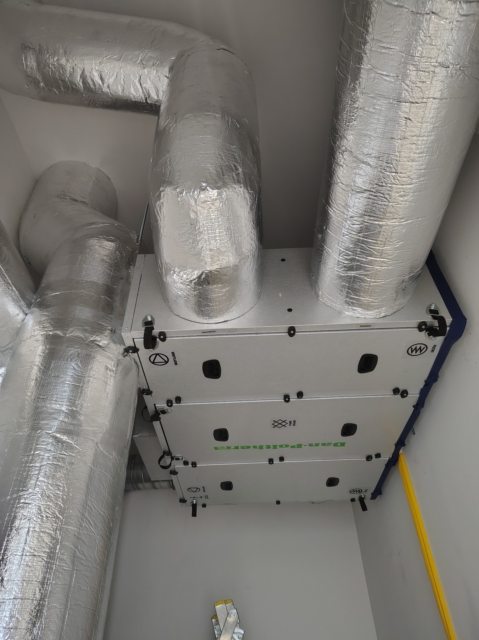 Zamontowany rekuperator Dan-Poltherm z izolowanymi przewodami wentylacyjnymi w pomieszczeniu, widok z dołu, żółta rurka instalacyjna w rogu.