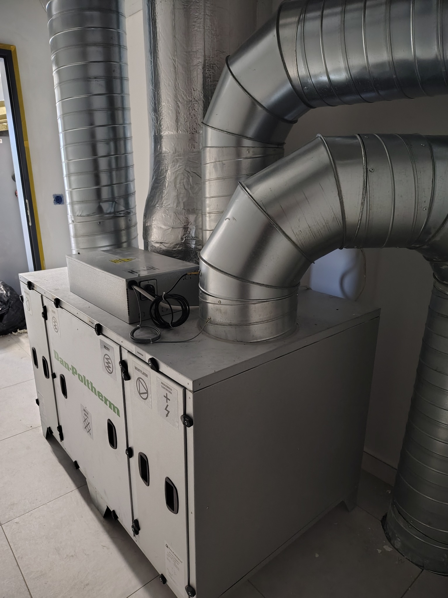 Szara centrala rekuperacyjna Dan-Poltherm z podłączonymi, metalowymi rurami wentylacyjnymi w pomieszczeniu. Widoczne naklejki informacyjne na obudowie.