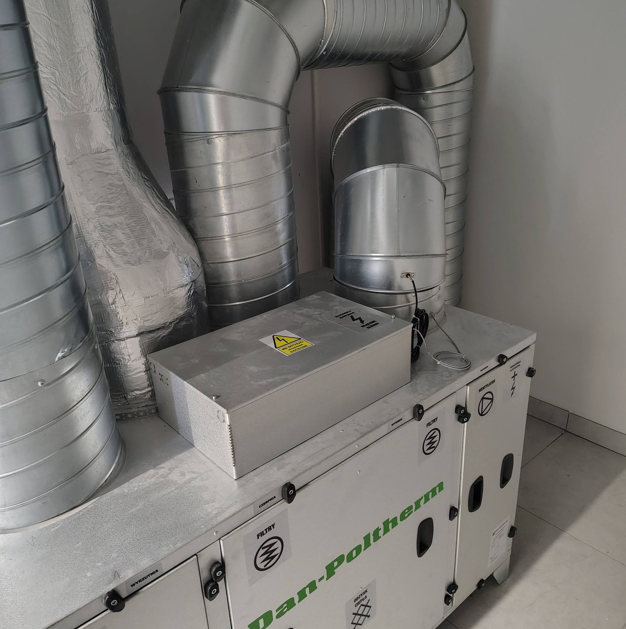 Centrala rekuperacyjna Dan-Poltherm z widocznymi filtrami i kanałami wentylacyjnymi. Na górze skrzynka elektryczna z ostrzeżeniem. Szare tło ściany i podłogi.