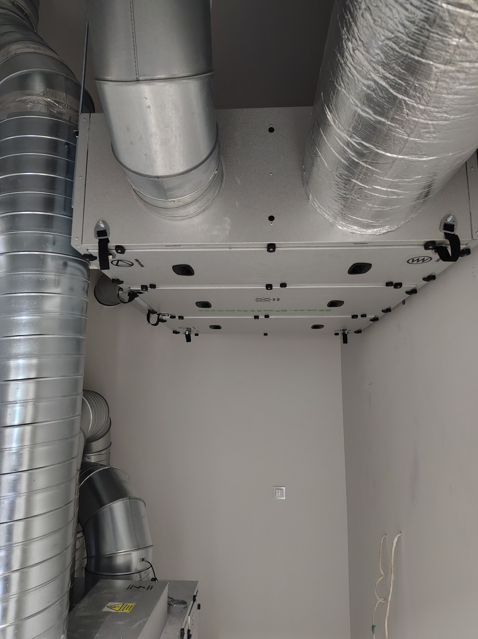 Zamontowana centrala rekuperacyjna z widocznymi przewodami wentylacyjnymi, srebrne rury i biała obudowa urządzenia na tle jasnej ściany.