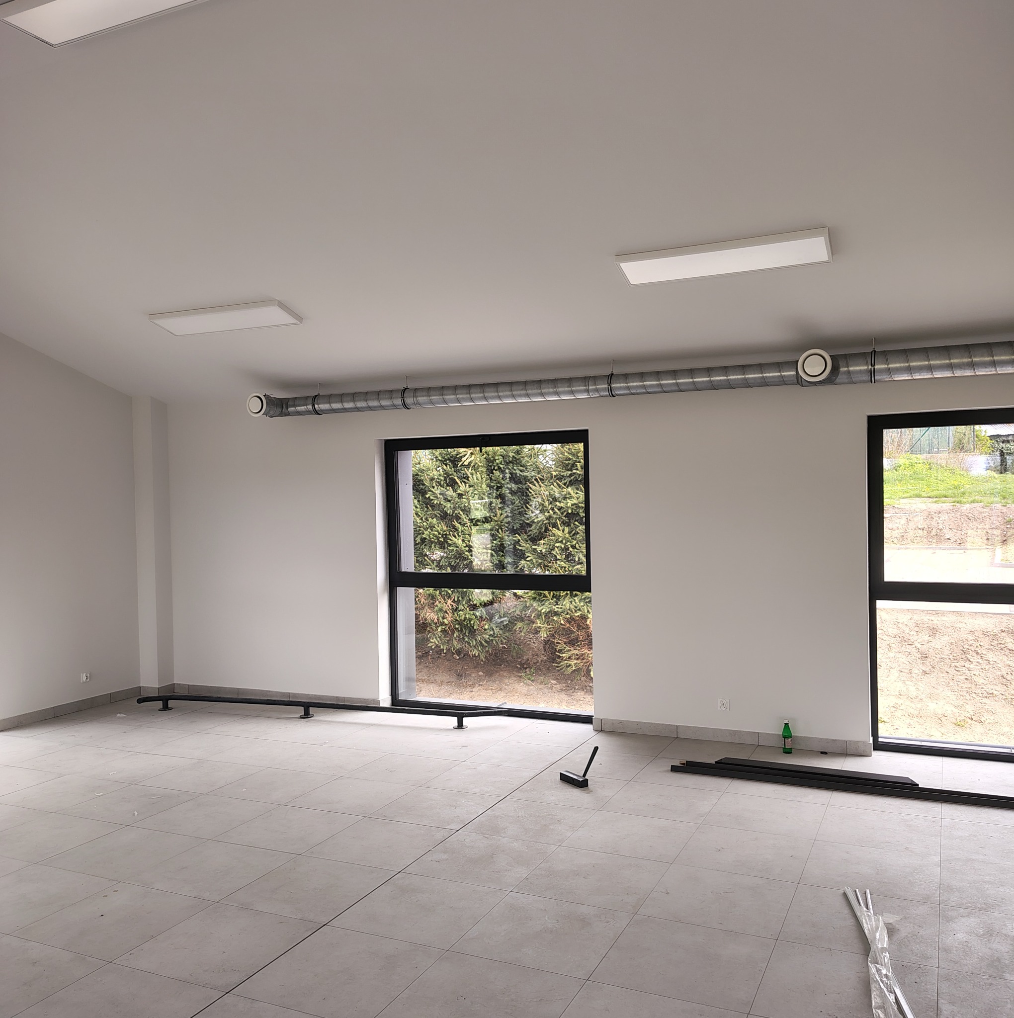 Wnętrze z szarą instalacją rekuperacji pod sufitem, widoczne okna z zielenią na zewnątrz. Na podłodze płytki i elementy instalacji. Oświetlenie sufitowe.
