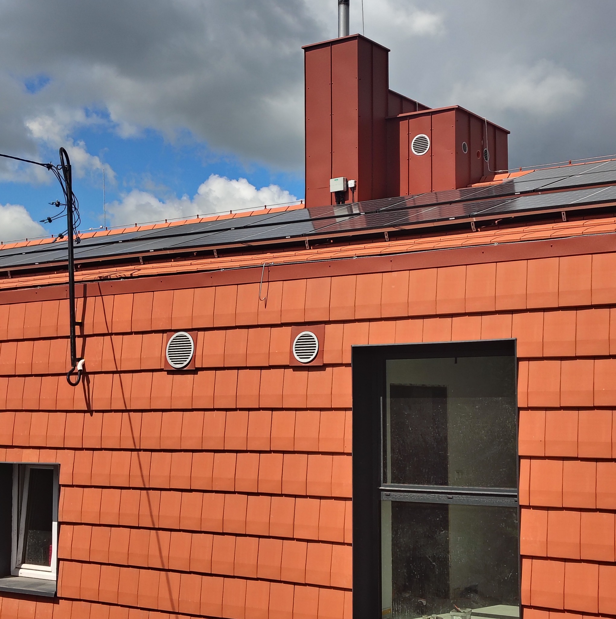 Czerwona elewacja budynku z panelami słonecznymi na dachu, kominem i kratkami wentylacyjnymi. Widoczne okno i instalacja elektryczna na tle pochmurnego nieba.
