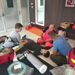 Instruktor w różowej kurtce demonstruje technikę resuscytacji krążeniowo-oddechowej na manekinie podczas kursu pierwszej pomocy, w tle widoczny sprzęt medyczny i drugi uczestnik kursu.