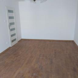 Stanisław Radchuk - Pusty pokój po remoncie z drewnianą podłogą, białe ściany i białe drzwi z szarymi paskami, wisi lampa z trzema białymi kloszami