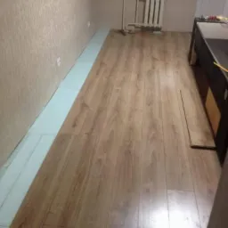 Proces układania paneli podłogowych w pokoju, widoczna podkładka izolacyjna przy ścianie oraz kilka niezamontowanych paneli.