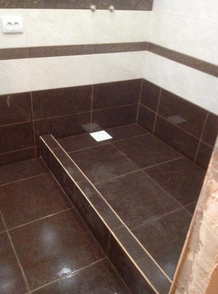 Wykończona kabina prysznicowa z brązowymi płytkami, dekoracyjnym pasem płytek i odpływem liniowym. Widoczne gniazdko elektryczne nad kabiną.