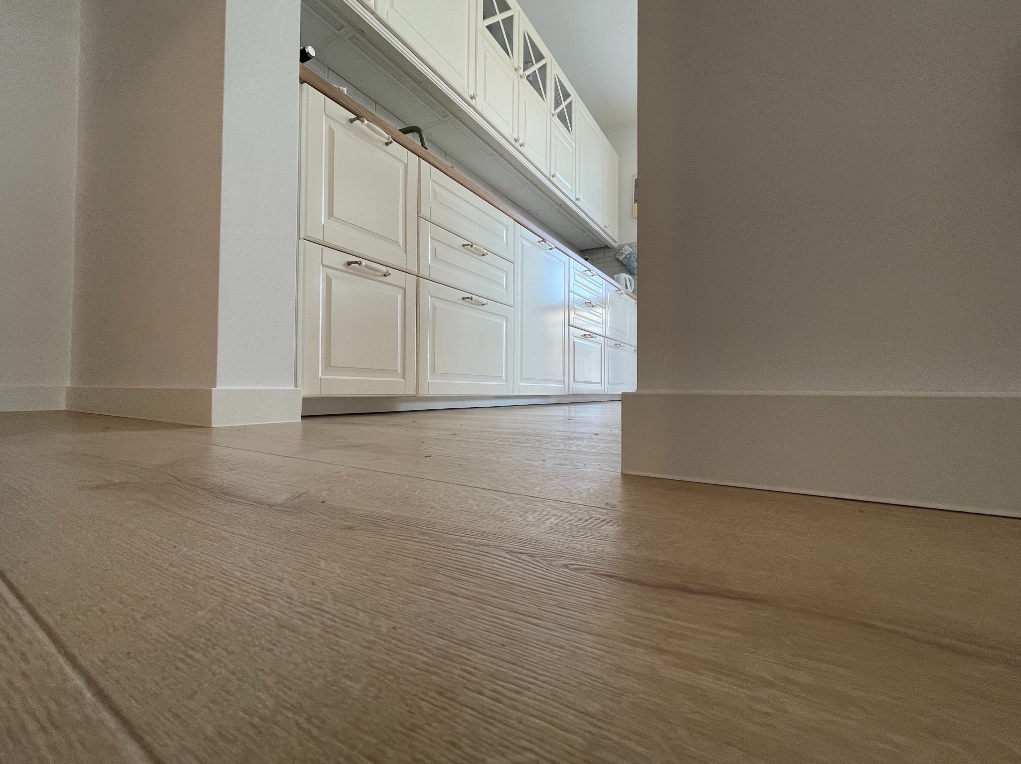 Jasne panele podłogowe w perspektywie z widoczną białą zabudową kuchenną w tle, z niskiego kąta.