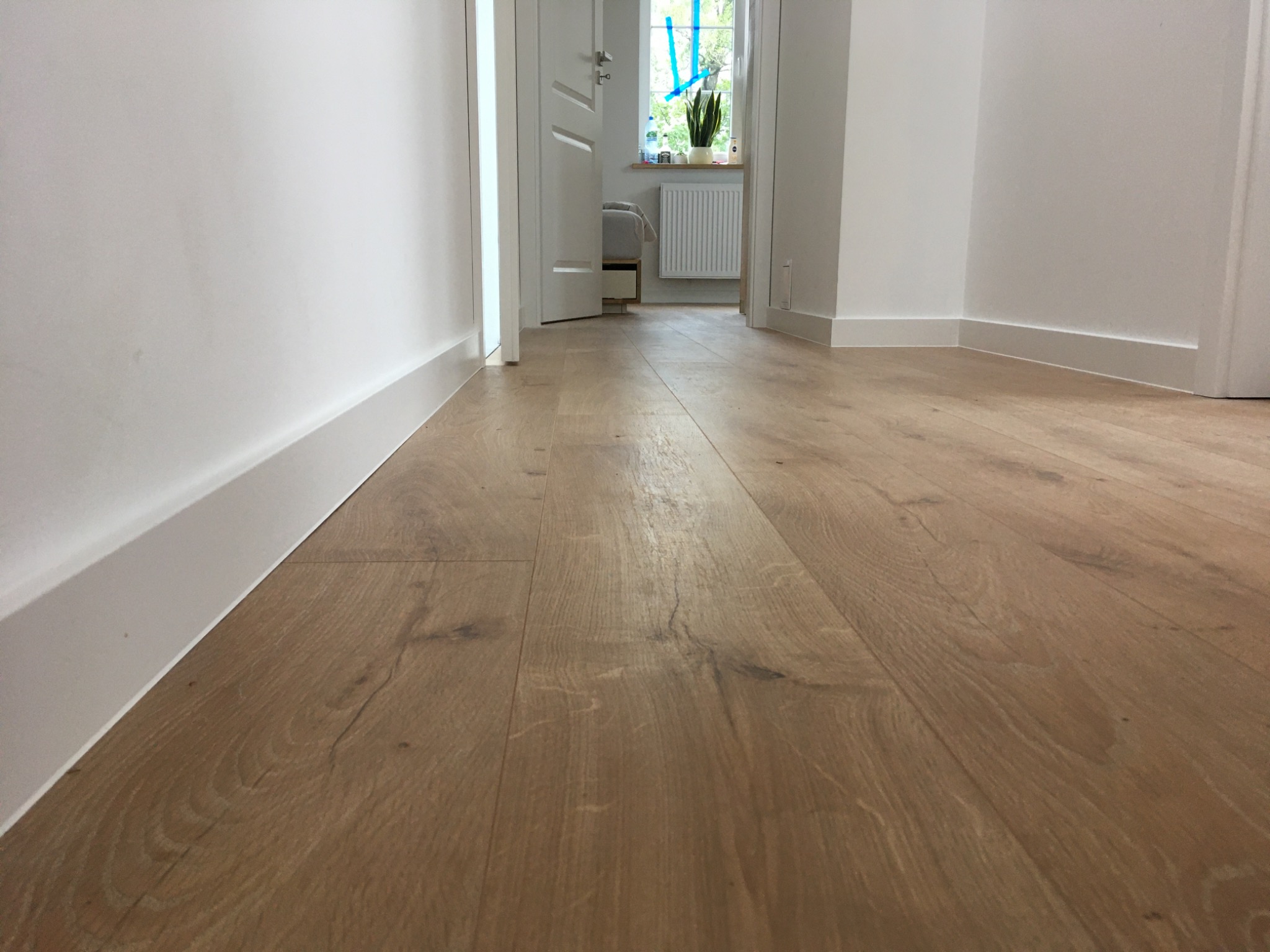 Jasna, drewniana podłoga w korytarzu z białymi listwami przypodłogowymi, widok w kierunku otwartych drzwi i okna z roślinami.