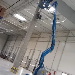 Pracownik w pomarańczowej kamizelce na podnośniku koszowym Genie Z-34/22N montuje system zabezpieczeń na słupie w hali magazynowej z widocznymi bramami segmentowymi.