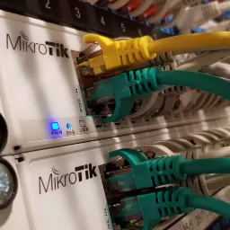 Dwa urządzenia MikroTik zamontowane w szafie rack, z podłączonymi kablami Ethernet w kolorach żółtym i zielonym, diody LED sygnalizują działanie.