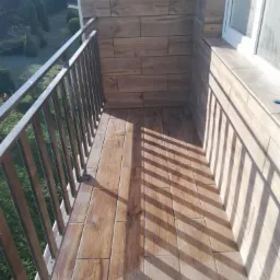 Wykończony balkon w Radomyślu Wielkim z podłogą i ścianami wyłożonymi płytkami imitującymi drewno, widoczne cienie rzucane przez metalową barierkę.