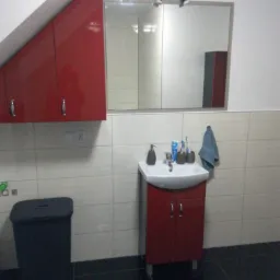 Ukośna szafka wisząca w kolorze czerwonym nad umywalką z szafką w łazience z czarną podłogą i lustrem z oświetleniem.