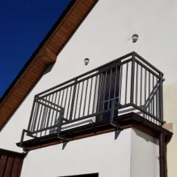 Czarna, metalowa balustrada balkonowa z prostymi szczeblami, zamontowana na białej elewacji domu w słoneczny dzień, widok z dołu.