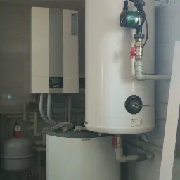 Kompaktowa kotłownia z dwoma zbiornikami: górnym z pompą obiegową i dolnym z szarą ściereczką na górze, oraz wiszącym kotłem gazowym Wolf, widoczne liczne rury i zawory.