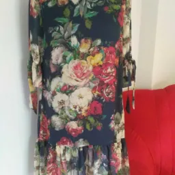 Sukienka z długim rękawem i kwiatowym wzorem, z falbaną na dole, prezentowana na manekinie na tle białej ściany i fragmentu czerwonej sofy.