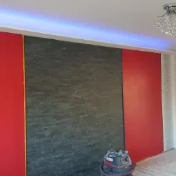 Wnętrze pokoju z czerwoną ścianą, fragmentem ściany wyłożonej szarym kamieniem dekoracyjnym i tapetą w srebrne pasy, podświetlony sufit LED, odkurzacz przemysłowy przy ścianie.