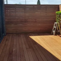 Nowoczesny taras z drewna egzotycznego, z drewnianą ścianą osłonową, aluminiowymi drzwiami przesuwnymi i srebrną drabinką opartą o krzew.