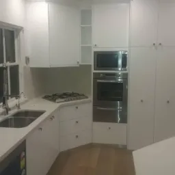 Nowoczesna, biała kuchnia z dużym oknem, widocznym zlewem, płytą grzewczą, piekarnikiem i szafkami. Minimalistyczny design, jasne drewniane podłogi.