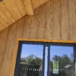 Fragment elewacji domu z oknem, imitacja drewna, widoczne odbicie otoczenia w szybie okiennej, zabezpieczenie na ramie okiennej.