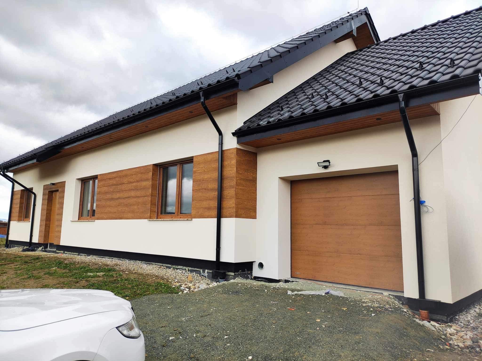 Nowoczesny dom jednorodzinny z elewacją w kolorze kremowym i panelami imitującymi drewno, czarną dachówką i orynnowaniem, oraz bramą garażową w kolorze drewna, widok z zewnątrz, pochmurne niebo.