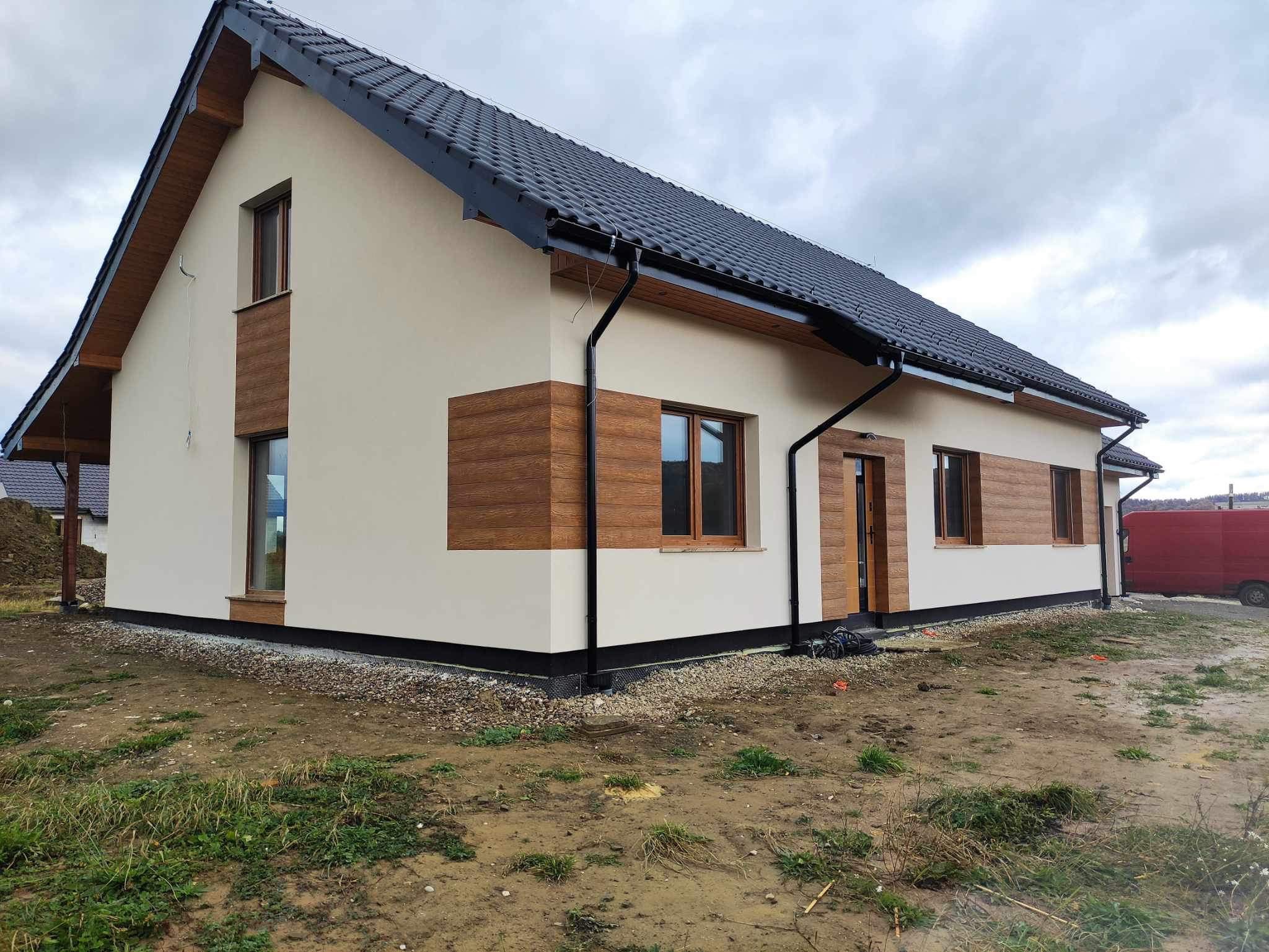 Nowoczesny dom jednorodzinny z jasną elewacją i drewnianymi panelami dekoracyjnymi, czarnym dachem i orynnowaniem, widok z zewnątrz na tle pochmurnego nieba.