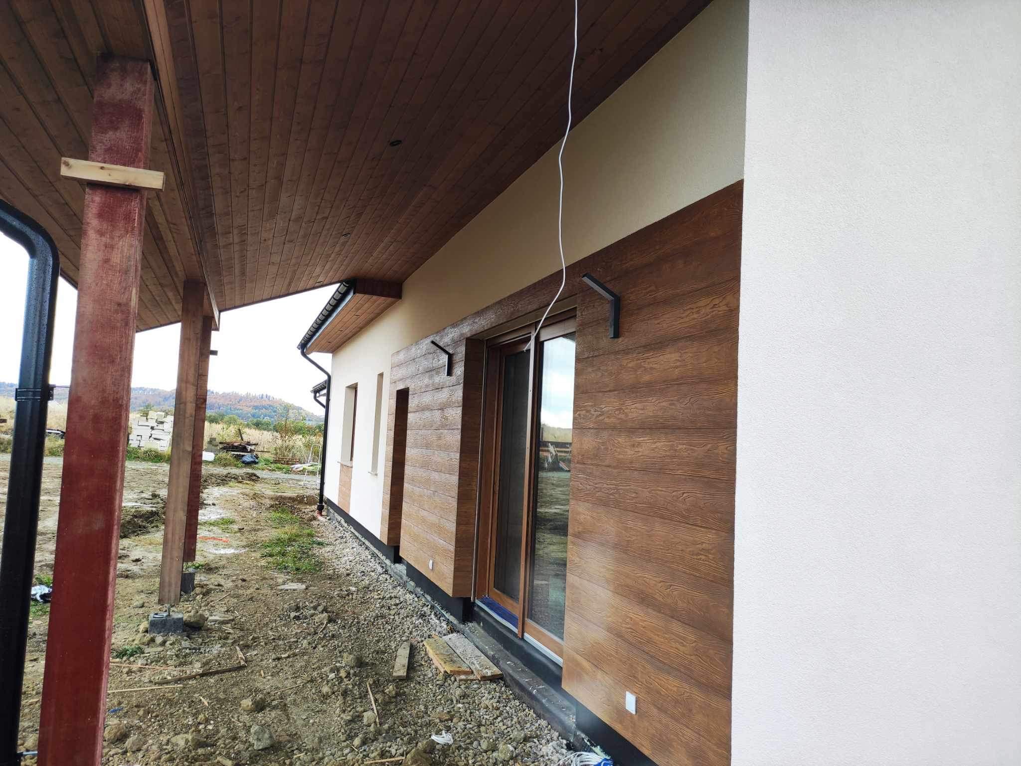 Fragment domu z drewnopodobną elewacją w kolorze orzecha włoskiego, z białym tynkiem strukturalnym po prawej stronie, widoczne okno tarasowe z brązowymi ramami i zadaszenie z drewnianą podbitką...