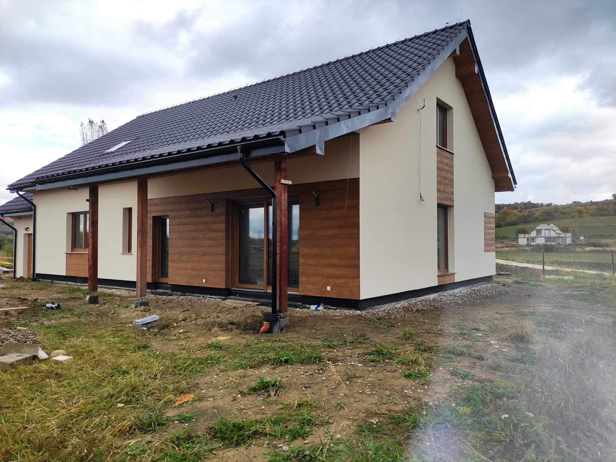 Nowoczesny dom jednorodzinny z beżową elewacją i drewnianymi panelami, czarną dachówką i podbitką, widok na otwarty teren i budowę w tle, pochmurne niebo.