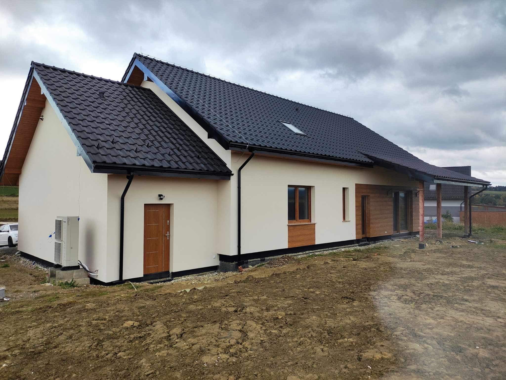Jednorodzinny dom z jasną elewacją, czarnym dachem z dachówki i drewnianymi elementami wykończeniowymi. Widoczny klimatyzator na ścianie i przygotowany teren wokół budynku.