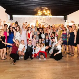 Duża grupa uśmiechniętych, elegancko ubranych osób pozuje do zdjęcia w jasnym wnętrzu z drewnianą podłogą i nowoczesnym oświetleniem, wszyscy machają rękami.