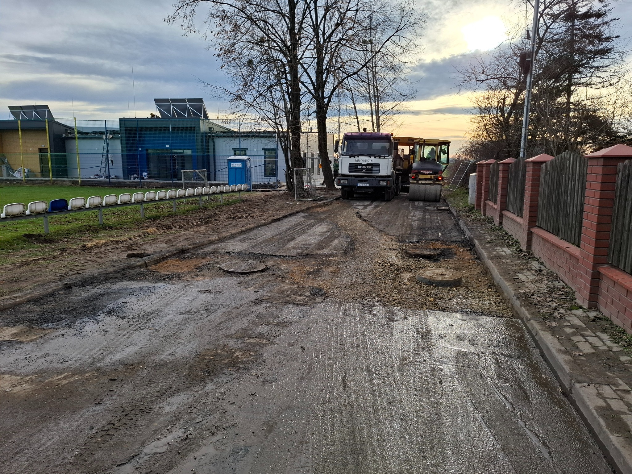 Nierówna, mokra droga w trakcie budowy lub remontu, widoczny walec drogowy i ciężarówka MAN z napisem 'Roboty Drogowe' na tle budynku sportowego ze panelami słonecznymi.