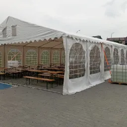 Rozłożony biały namiot z oknami, wewnątrz drewniane stoły i ławki. Na zewnątrz zbiorniki z wodą. Widok na szarą kostkę brukową i budynki w tle.